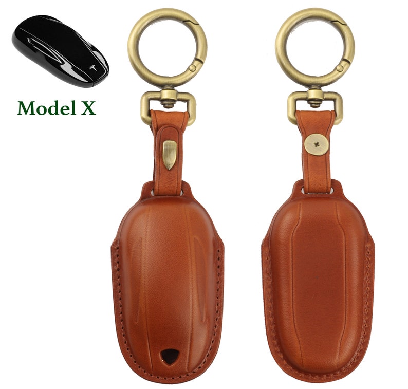 May include: A brown leather key chain with a gold metal key ring and a gold metal clasp. The key chain is designed to hold a Tesla Model X key fob. The key chain is shown next to a black Tesla Model X key fob. The text "Model X" is shown in green letters.