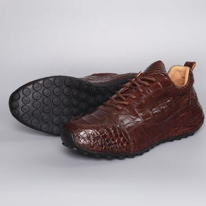 Puede incluir: Zapatillas de cuero marr&oacute;n con textura de piel de cocodrilo. Los zapatos tienen punta redondeada, cierre de cordones y suela de goma negra. Estas zapatillas informales tienen un aspecto texturizado y estampado.