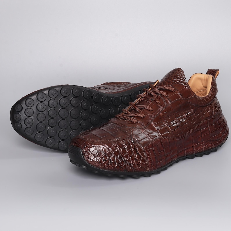 Puede incluir: Zapatillas de cuero marr&oacute;n con textura de piel de cocodrilo. Los zapatos tienen punta redondeada, cierre de cordones y suela de goma negra. Estas zapatillas informales tienen un aspecto texturizado y estampado.