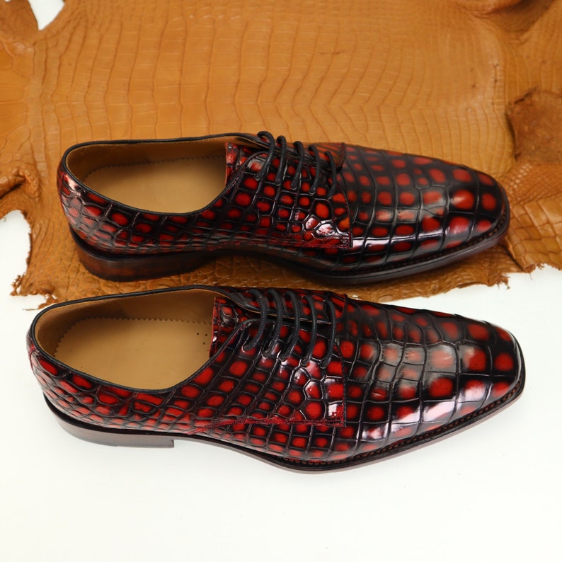 May include: A pair of red leather dress shoes with a crocodile pattern. The shoes have black laces and a pointed toe.