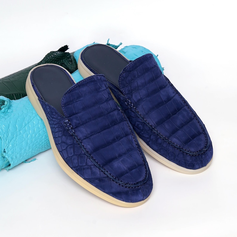 May include: A pair of navy blue leather slip-on shoes with a crocodile texture. The shoes have a cream-colored sole and a dark blue interior. The shoes are displayed on a white surface with green and blue leather scraps in the background.