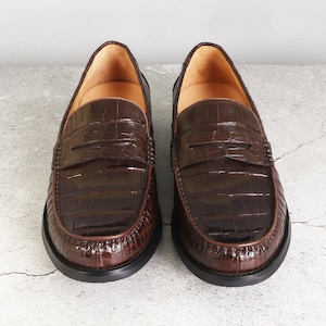 Puede incluir: Un par de mocasines de cuero marr&oacute;n con estampado de cocodrilo. Los mocasines tienen un dise&ntilde;o de deslizamiento y una punta redonda.