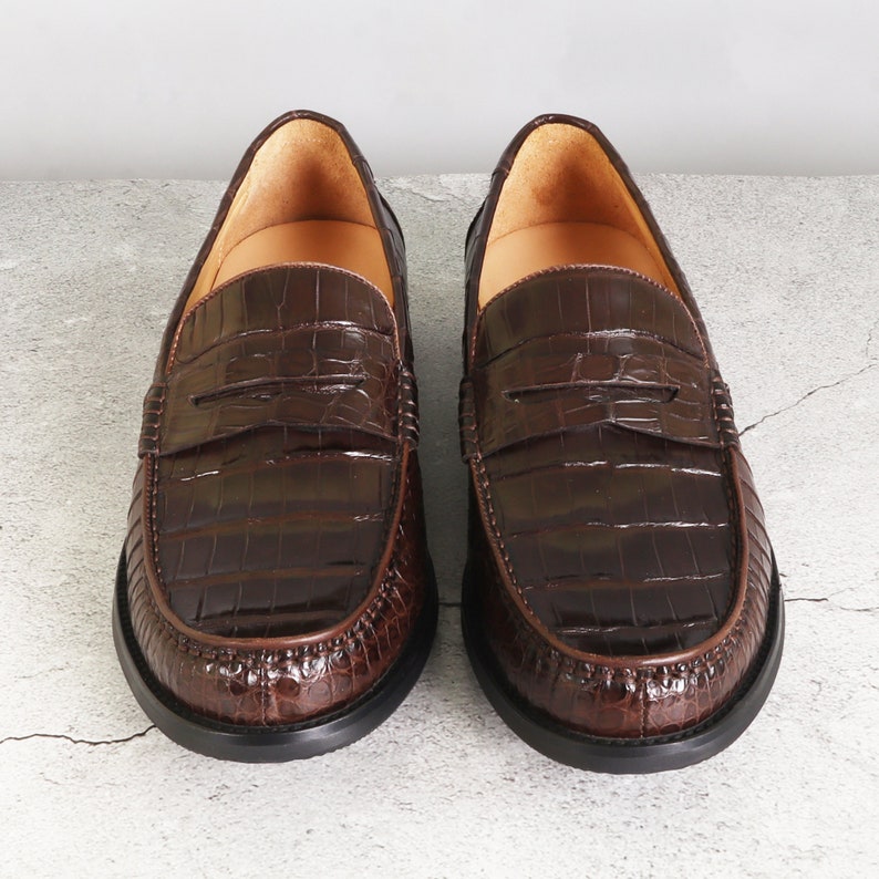 Puede incluir: Un par de mocasines de cuero marr&oacute;n con estampado de cocodrilo. Los mocasines tienen un dise&ntilde;o de deslizamiento y una punta redonda.