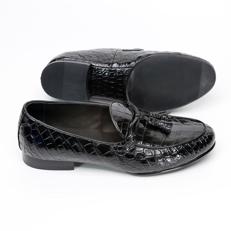 May include: Black leather loafers with a crocodile skin texture. The shoes feature a tassel detail and a low heel. One shoe is positioned above the other, showcasing the sole and the overall design. The shoes are on a white background.