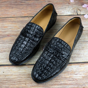 May include: A pair of black leather loafers with a crocodile skin texture. The shoes have a classic slip-on design with a low heel and a light brown interior. The loafers are displayed on a wooden surface.