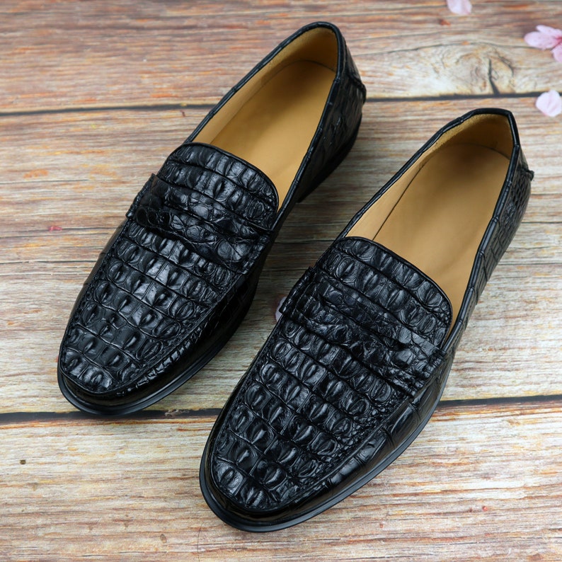 May include: A pair of black leather loafers with a crocodile skin texture. The shoes have a classic slip-on design with a low heel and a light brown interior. The loafers are displayed on a wooden surface.