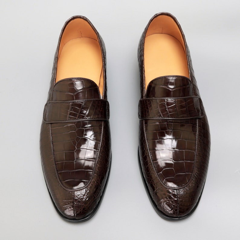 May include: A pair of brown leather loafers with a crocodile pattern. The loafers have a pointed toe and a slip-on design.