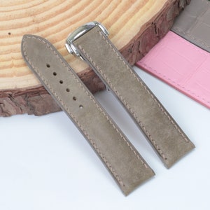 Puede incluir: Dos correas de reloj de cuero gris con costuras blancas. Las correas están dispuestas en plano sobre una superficie de madera.