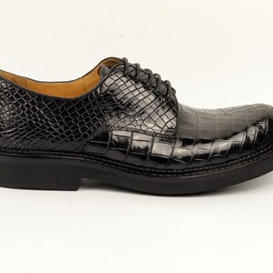 May include: Black leather dress shoes with a crocodile pattern. The shoes have a lace-up closure and a thick sole.