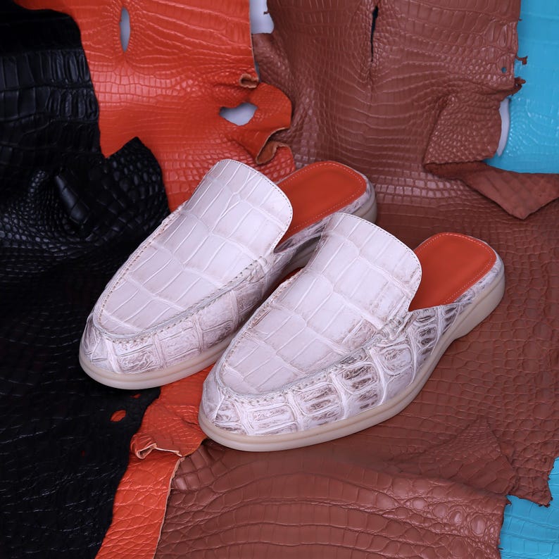 May include: A pair of white crocodile-textured leather slip-on shoes with orange inner soles. The shoes are displayed on a background of various colored leather hides, including black, orange, brown, and blue.