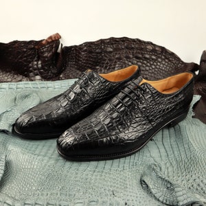 Op de afbeelding: Een paar zwarte leren veterschoenen met een krokodilpatroon. De schoenen liggen op een lichtblauwe en bruine alligatorhuid achtergrond.