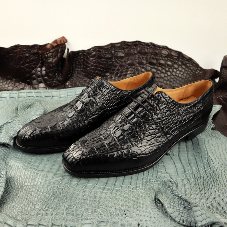 Op de afbeelding: Een paar zwarte leren veterschoenen met een krokodilpatroon. De schoenen liggen op een lichtblauwe en bruine alligatorhuid achtergrond.