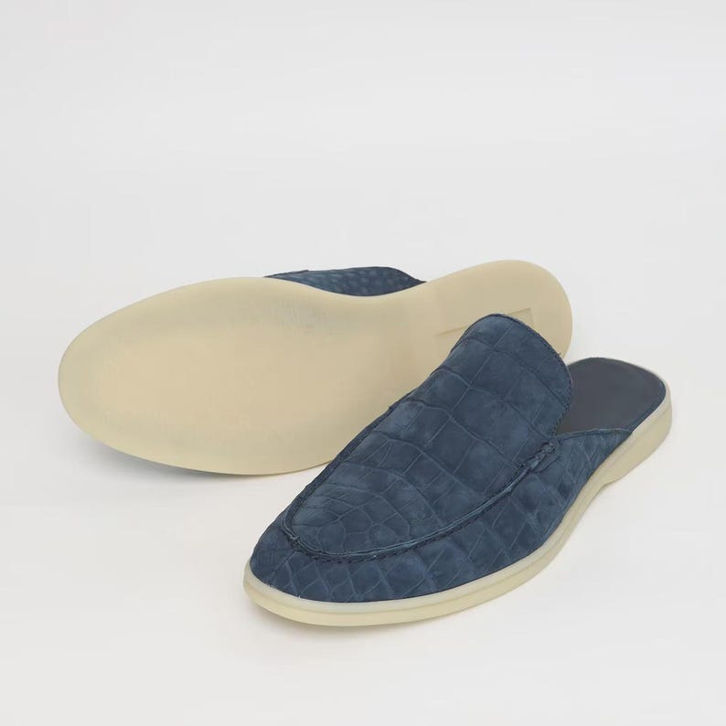 May include: A pair of blue suede slip-on shoes with a crocodile pattern. The shoes have a light-colored sole and a rounded toe. One shoe is upside down, revealing the sole.