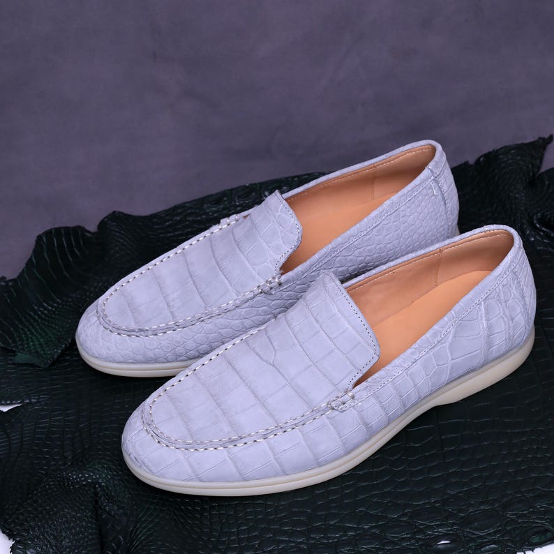 May include: A pair of light blue leather loafers with a crocodile skin texture. The shoes have a slip-on design with a stitched seam around the vamp and a tan interior. The soles are off-white.
