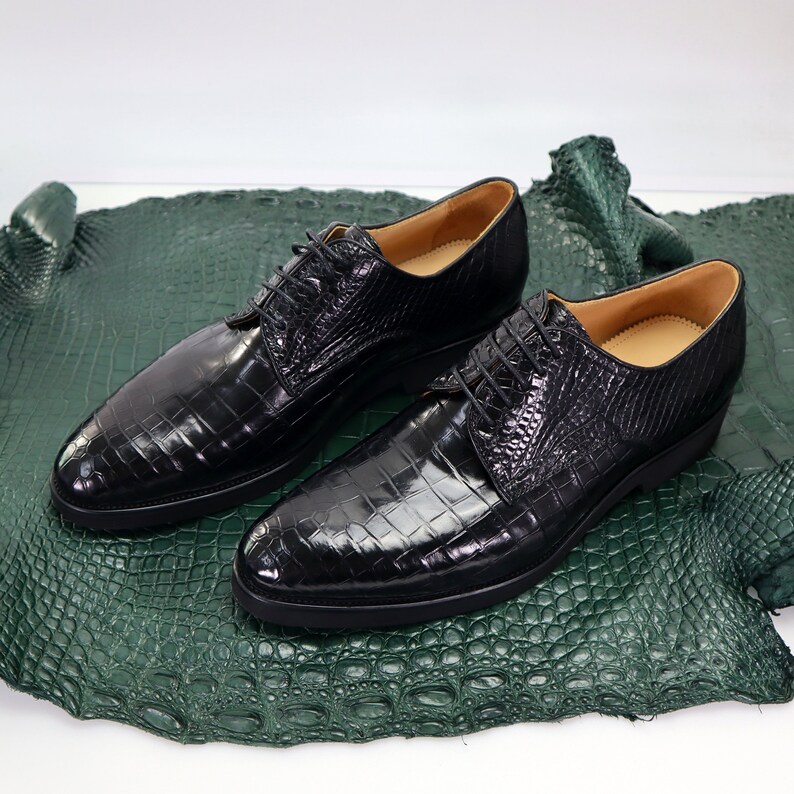 K&ouml;nnte beinhalten: Ein Paar schwarze Lederschuhe mit Kroko-Pr&auml;gung. Die Schuhe haben Schn&uuml;rsenkel und liegen auf einer gr&uuml;nen Krokodilleder-Oberfl&auml;che.