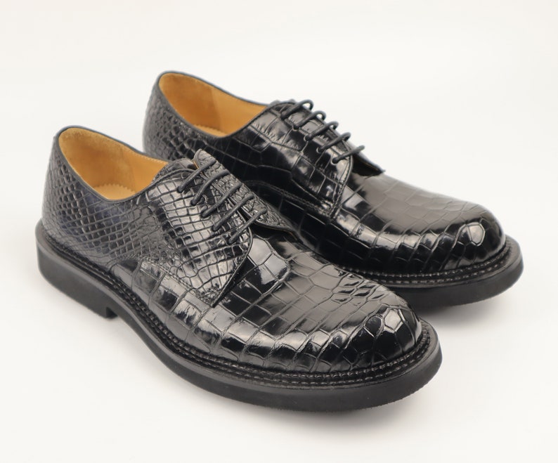 Pair of black Alligator dress shoes displayed side by side on a wooden surface, showcasing lace-up design, pointed toes, and glossy finish with visible scale texture.