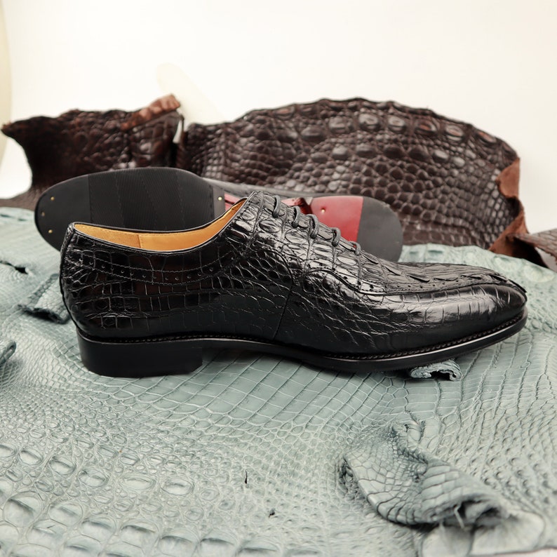 Op de afbeelding: Zwarte leren herenschoenen met een krokodillenpatroon. De schoenen liggen op een stuk alligatorleer.