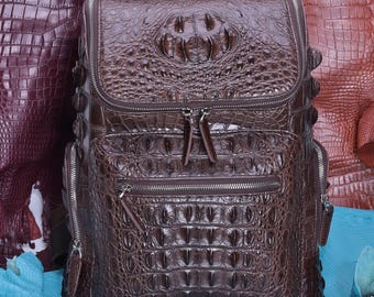 Pode incluir: Uma mochila castanho-escura com um padrão de textura de pele de crocodilo. A mala inclui uma aba superior com fecho de correr, um bolso frontal com fecho e compartimentos laterais com fecho. As ferragens são metálicas.