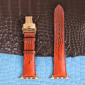 Pode incluir: Duas pulseiras de relógio de couro laranja com ferragens douradas. As pulseiras são mostradas em um fundo de padrão de crocodilo marrom e azul.