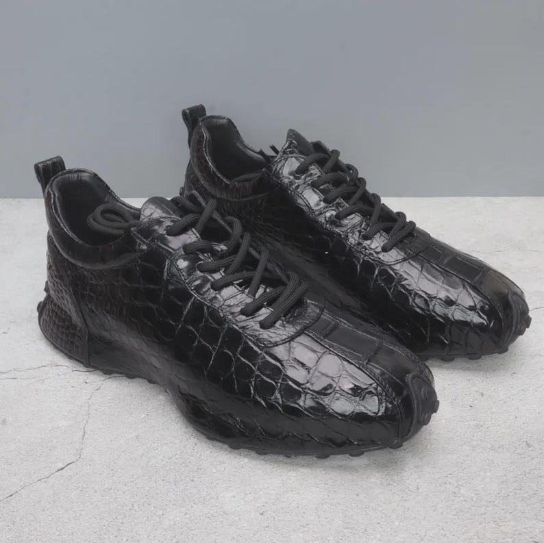 Puede incluir: Un par de zapatillas negras con un patr&oacute;n texturizado similar al de la piel de cocodrilo. Los zapatos tienen cordones negros y una suela gruesa negra. Las zapatillas est&aacute;n dise&ntilde;adas para uso casual.