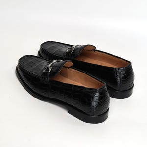 Op de afbeelding: Een paar zwarte leren loafers met een krokodillenstructuur. De schoenen hebben een zilverkleurige gesp en een ronde neus. De binnenkant van de schoen is bruin.