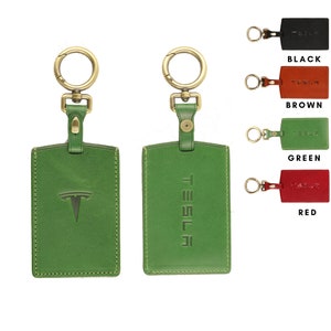 May include: Green leather Tesla key chain with a gold metal ring and a Tesla logo on the front. The keychain is shown next to three other keychains in black, brown, and red leather.