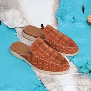 May include: A pair of brown leather slip-on shoes with a crocodile pattern. The shoes have a light tan sole and a brown interior. The shoes are displayed on a blue and white textured surface.