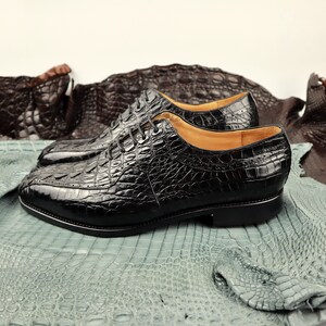 Op de afbeelding: Zwarte leren herenschoenen met een krokodillenhuidpatroon. De schoenen liggen op een lichtblauw alligatorhuid oppervlak.