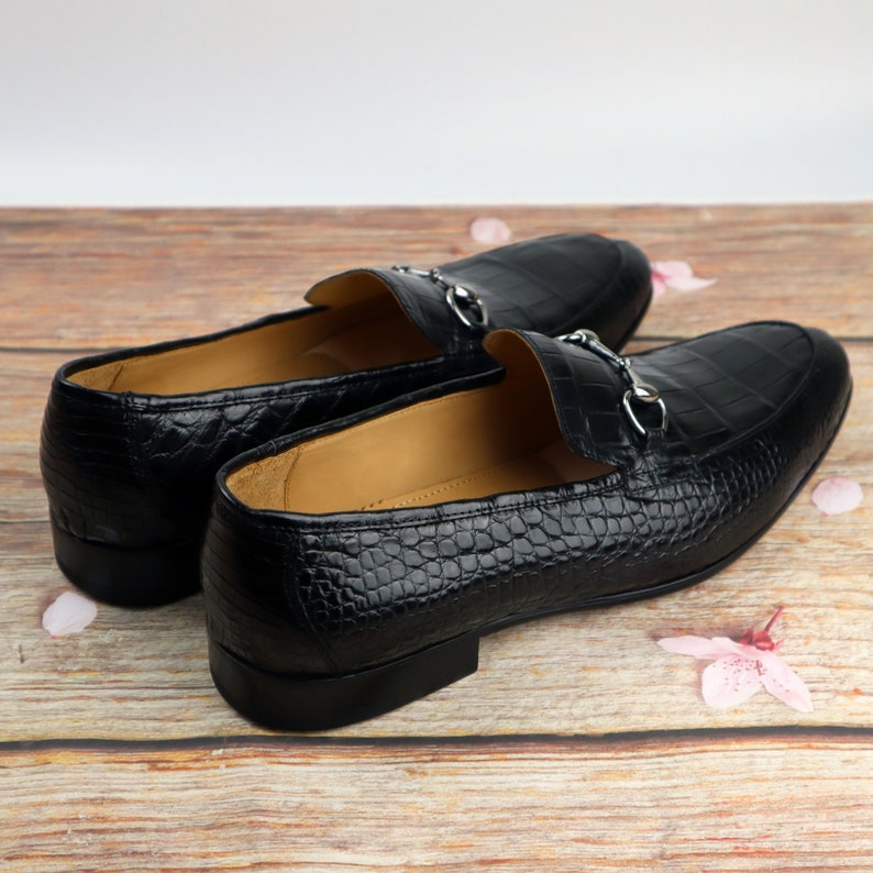May include: A pair of black leather loafers with a crocodile pattern and a silver buckle detail. The loafers are on a wooden surface with pink flower petals.