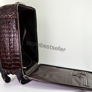 May include: A brown crocodile-embossed leather suitcase with a zippered pocket on the front and a handle on the top. The suitcase is open and the interior is lined with brown fabric. The suitcase has four wheels and a retractable handle.