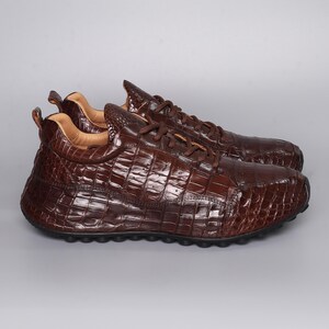 Puede incluir: Un par de zapatillas de cuero marr&oacute;n con textura de piel de cocodrilo. Los zapatos tienen cordones y una puntera redondeada. Las suelas son negras y el interior es de un marr&oacute;n m&aacute;s claro. Las zapatillas se muestran sobre un fondo gris.