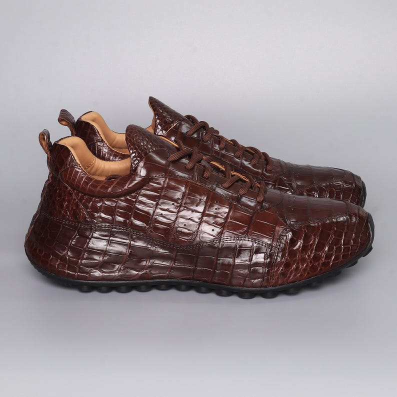 Puede incluir: Un par de zapatillas de cuero marr&oacute;n con textura de piel de cocodrilo. Los zapatos tienen cordones y una puntera redondeada. Las suelas son negras y el interior es de un marr&oacute;n m&aacute;s claro. Las zapatillas se muestran sobre un fondo gris.