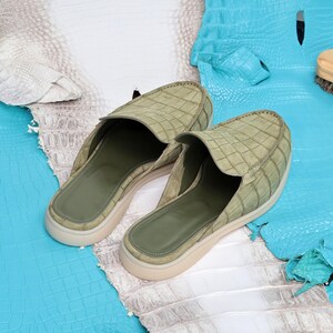 May include: A pair of sage green leather slip-on shoes with a textured, crocodile-like pattern. The shoes have a rounded toe and a low, off-white sole. The interior is a darker green. The shoes are displayed on a backdrop of blue and white leather.