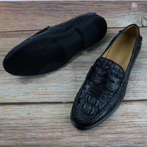 May include: Black leather loafers with a crocodile skin texture. One shoe is upright, showcasing the textured vamp and toe, while the other is inverted, revealing the sole. The shoes are set against a wooden background.