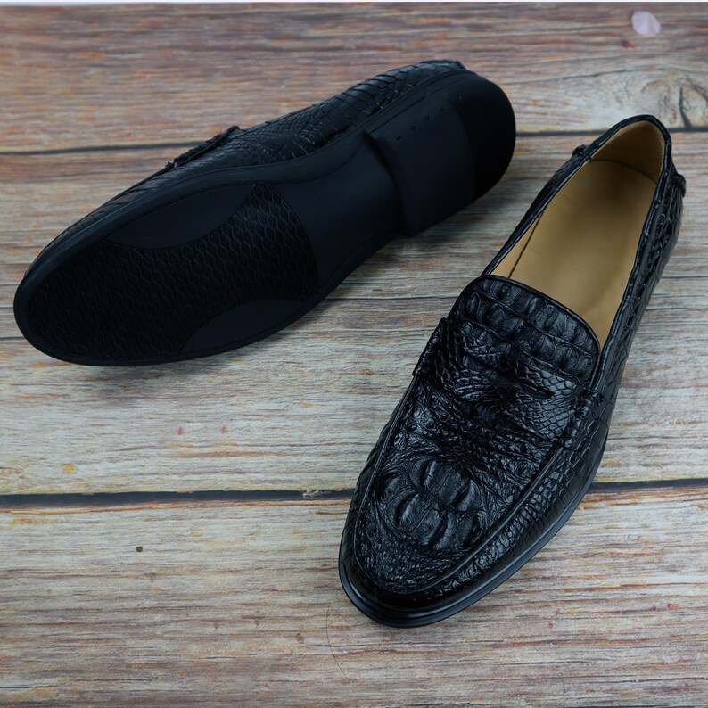 May include: Black leather loafers with a crocodile skin texture. One shoe is upright, showcasing the textured vamp and toe, while the other is inverted, revealing the sole. The shoes are set against a wooden background.