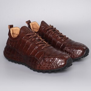Puede incluir: Un par de zapatillas de cuero marr&oacute;n con textura de piel de cocodrilo. Los zapatos tienen cordones marrones, una leng&uuml;eta acolchada y una suela negra. Las zapatillas se muestran sobre un fondo gris claro.