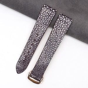 Puede incluir: Dos correas de reloj con un patrón texturizado y moteado en tonos grises y blancos. Las correas tienen un color base gris oscuro con marcas circulares más claras. Una correa presenta una hebilla dorada.