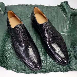 K&ouml;nnte beinhalten: Ein Paar schwarze Lederschn&uuml;rschuhe mit Kroko-Muster. Die Schuhe liegen auf einem gr&uuml;nen Krokodilleder-Hintergrund.