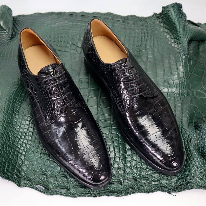 K&ouml;nnte beinhalten: Ein Paar schwarze Lederschn&uuml;rschuhe mit Kroko-Muster. Die Schuhe liegen auf einem gr&uuml;nen Krokodilleder-Hintergrund.