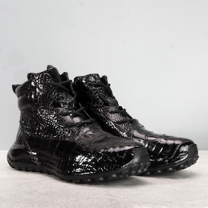 Może przedstawiać: Para czarnych skórzanych butów sportowych o wysokim profilu z tłoczonym wzorem krokodyla. Buty mają grubą podeszwę i sznurowanie.