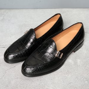 Könnte beinhalten: Ein Paar schwarze Leder-Loafer mit Krokodiltextur. Die Schuhe haben eine goldfarbene Schnalle und ein Lederinnenfutter. Die Sohlen sind schwarz mit einem roten Akzent.