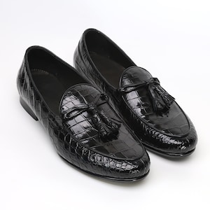 May include: A pair of black leather loafers with a glossy, textured finish. Each shoe features a decorative tassel and a rounded toe. The shoes are displayed on a white background, highlighting their polished appearance.