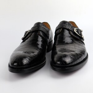 May include: A pair of black leather shoes with a crocodile pattern. The shoes have a buckle fastening and a pointed toe.