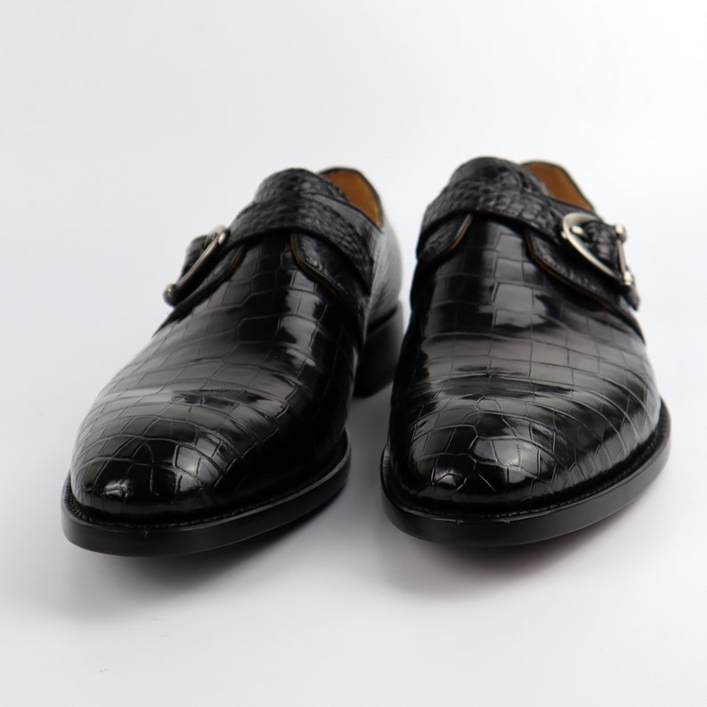 May include: A pair of black leather shoes with a crocodile pattern. The shoes have a buckle fastening and a pointed toe.