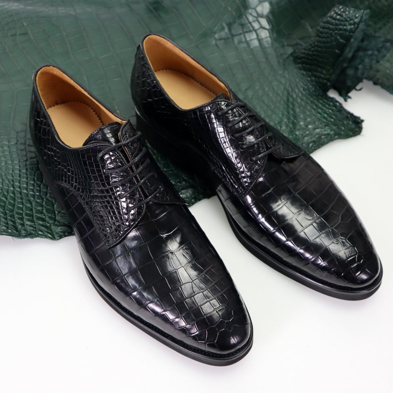 K&ouml;nnte beinhalten: Ein Paar schwarze Lederschuhe mit Kroko-Muster. Die Schuhe haben eine Schn&uuml;rung vorne und eine spitze Zehenpartie.