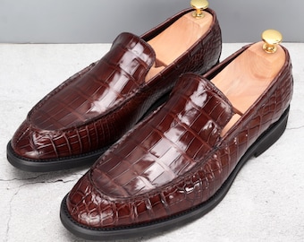 Peut inclure: Une paire de mocassins en cuir marron avec un motif peau de crocodile. Les chaussures ont un bout arrondi et une semelle noire. Des embauchoirs en bois avec des boutons dorés sont insérés dans chaque chaussure.