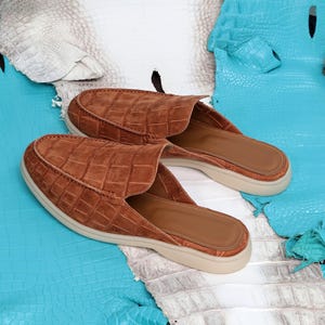 May include: A pair of brown leather slip-on shoes with a textured, crocodile-like pattern. The shoes have a rounded toe and a beige sole. The background features blue and white leather materials.