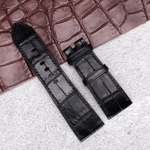 May include: A black watch strap featuring a textured, embossed pattern that mimics the appearance of alligator skin. The strap consists of two separate pieces, with the longer section containing multiple adjustment holes and the shorter section.
