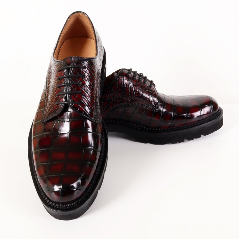 May include: A pair of dark red leather shoes with a crocodile pattern. The shoes have laces and a rounded toe.