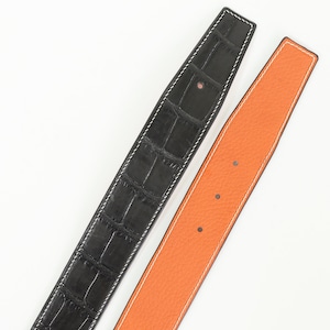 以下が含まれることがあります： 黒いワニ革風の質感と白いステッチが施されたベルトと、オレンジ色の滑らかな質感と白いステッチが施された2本の革ベルト。 白い背景に表示され、対照的な色と質感を際立たせています。